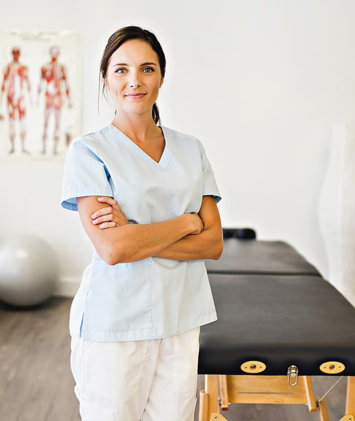 A confident female healthcare professional in scrubs stands with arms crossed in a treatment room.