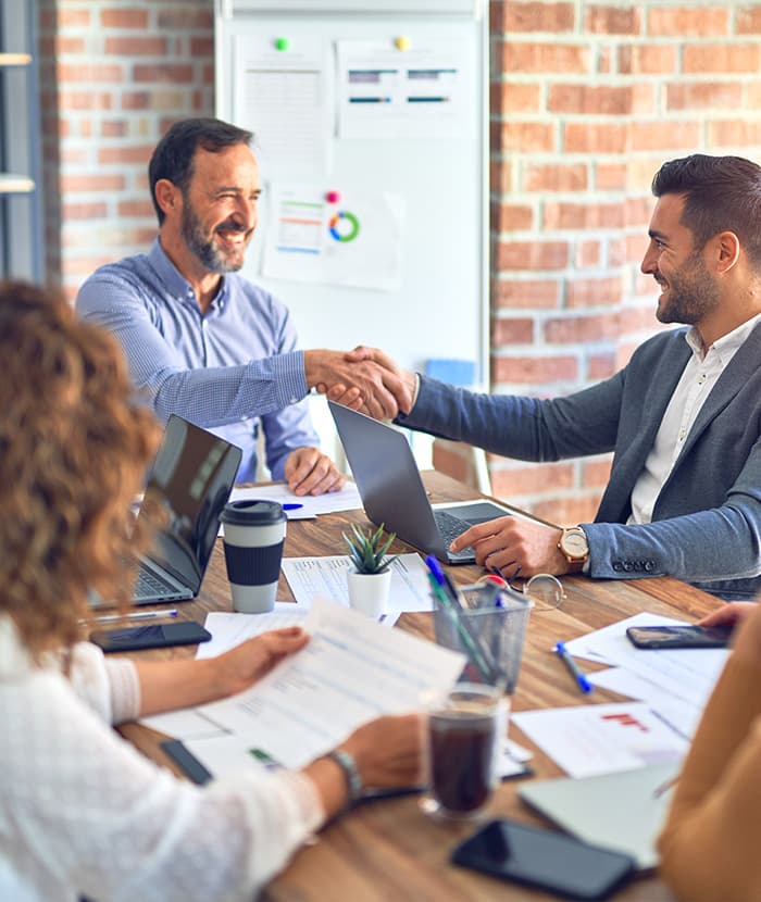 Two men shake hands at a conference table filled with laptops and paperwork, symbolizing professional collaboration.