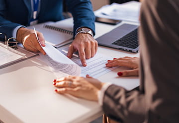 A person points to a document while discussing it with another person at a desk, suggesting a legal consultation.
